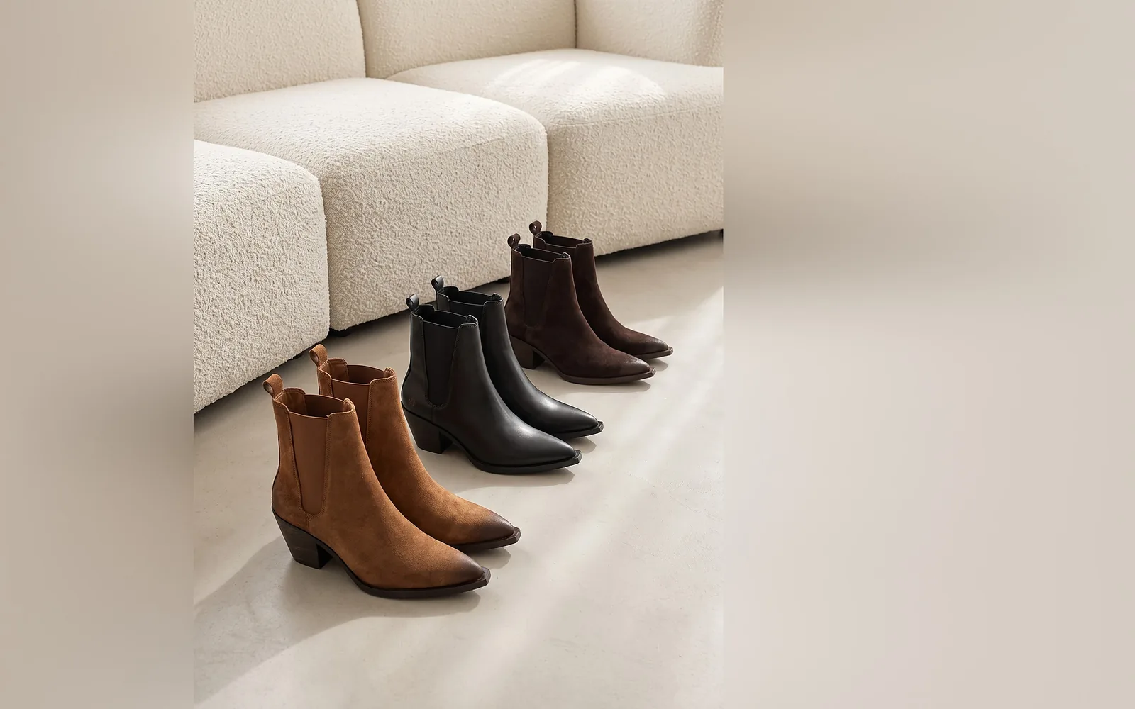 Drei spitze Chelsea Boots von Apple of Eden in Cognac, Schwarz und Dunkelbraun vor einem hellen Bouclé-Sofa